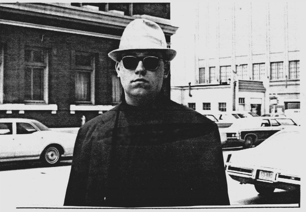 Photo of a man in black attire with a white fedora hat and dark sunglasses staring at the camera.