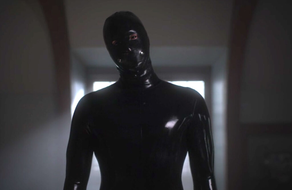 A human looking figure in an all black rubber suit stares at the camera.